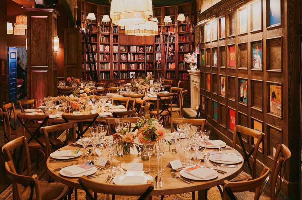 Tables set for a private dinner, surrounded by built-in bookshelves, floral centerpieces, and soft, ambient lighting.