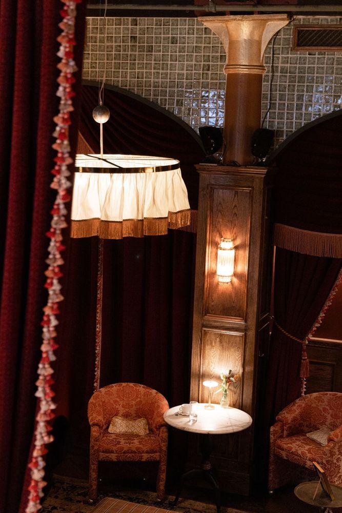 Visual Comfort fixtures including fringed lampshades and sconces create warm, private dining spaces framed by elegant curtains and marble-topped tables.