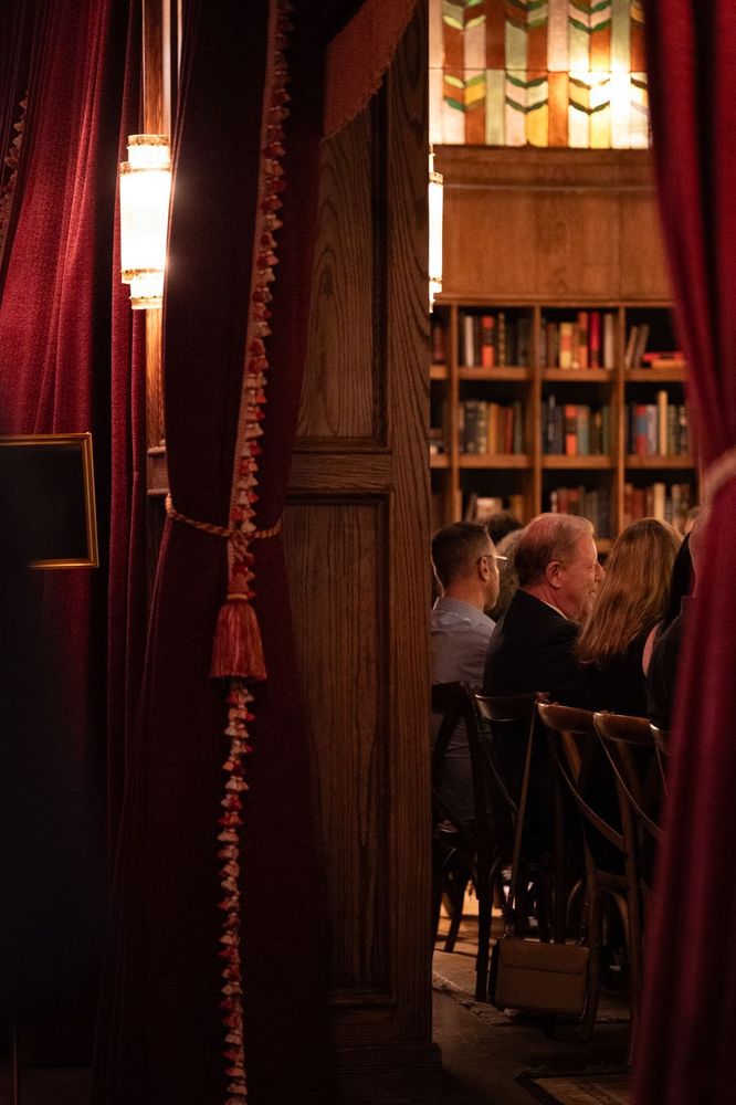 Curtains trimmed with tassels frame a view of seated guests beneath soft wall-mounted lighting.