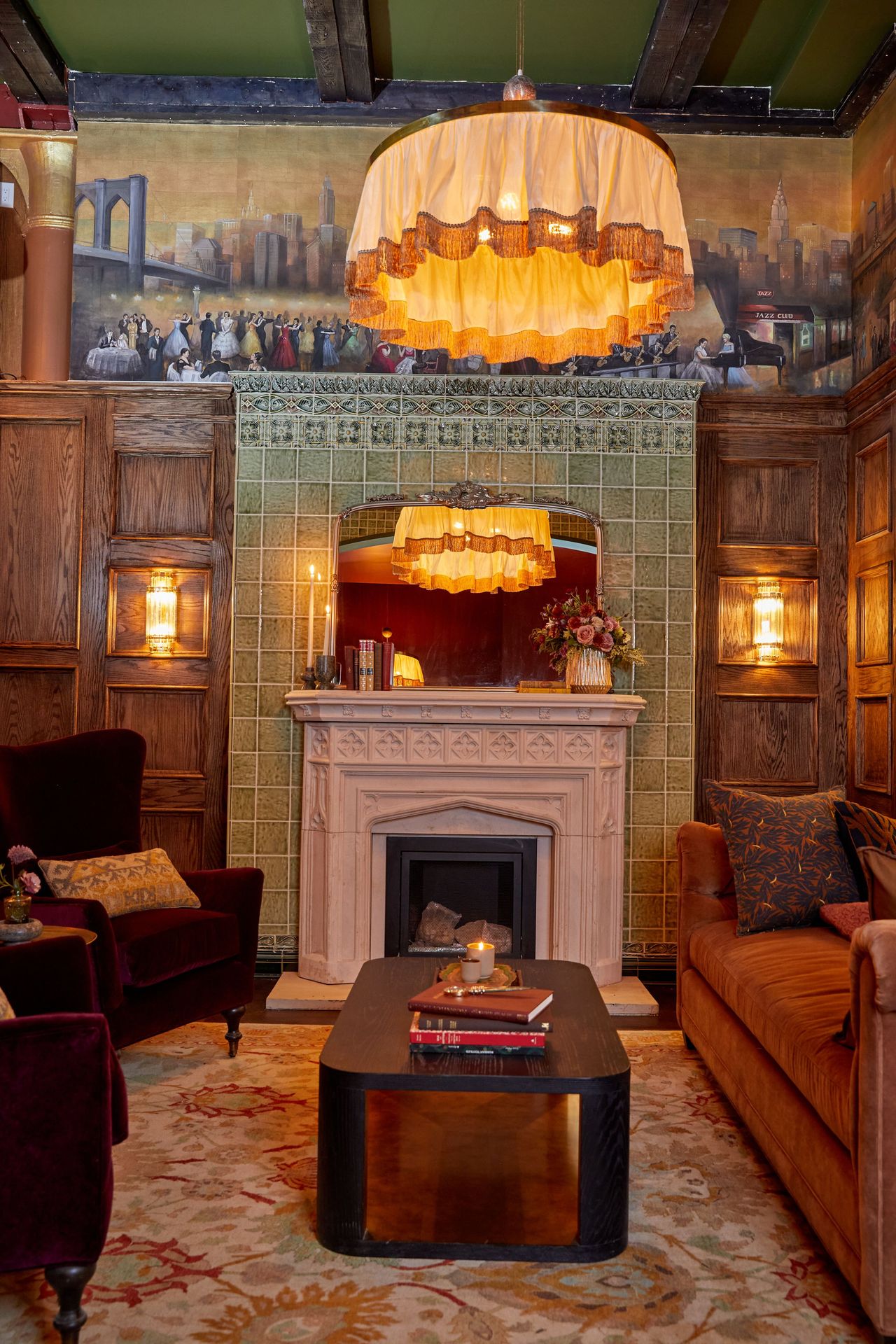 A carved stone fireplace sits beneath a mural of a city skyline, framed by green tilework by Pratt & Larson in Maxwell's Grand Room